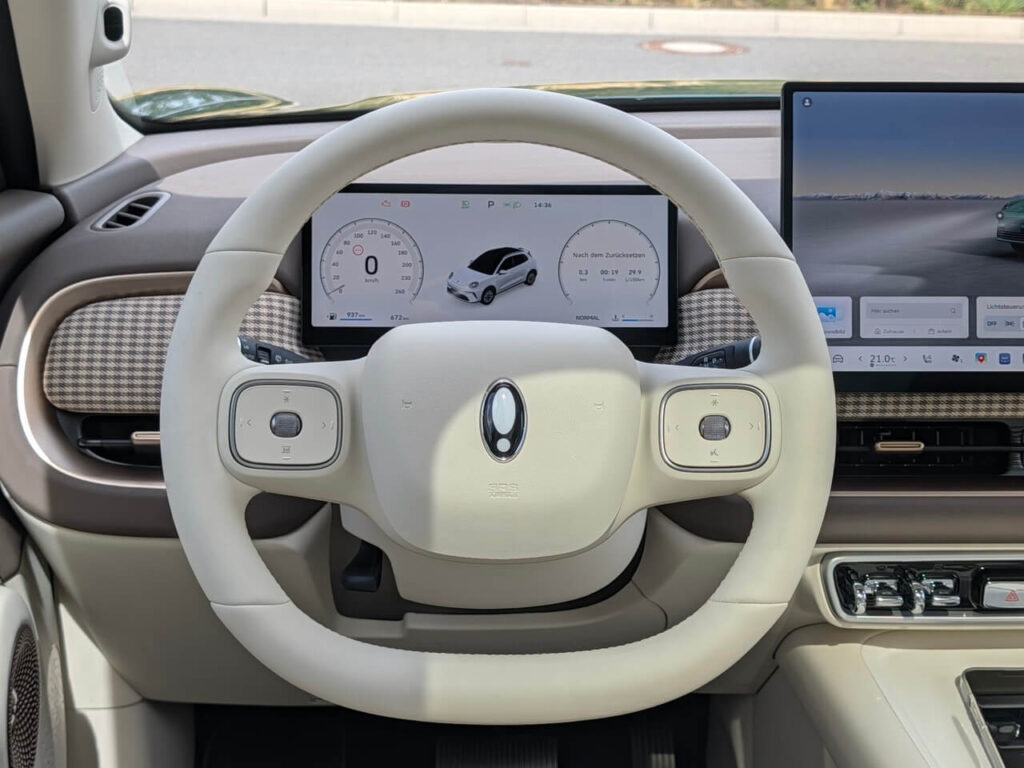 Fahrerperspektive im GWM Ora 5 Luxury: Weich gepolstertes 2-Speichen-Lenkrad mit hellem Kunstleder und praktischen Multifunktionstasten. Das digitale Kombiinstrument lässt sich individuell anpassen.